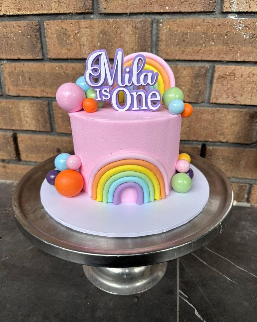 Pink rainbow birthday cake for Mila is One with pastel rainbow fondant arch, colourful ball toppers and Mila is One acrylic topper