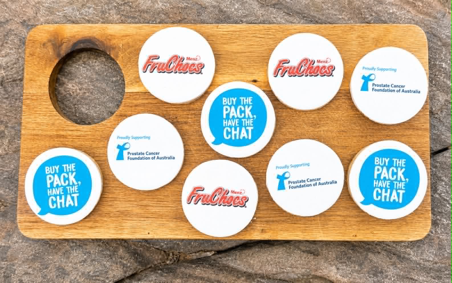 Round white sugar cookies on a wooden board — FruChocs Menz logo, blue Buy the Pack Have the Chat speech bubble, and Prostate Cancer Foundation of Australia Proudly Supporting designs