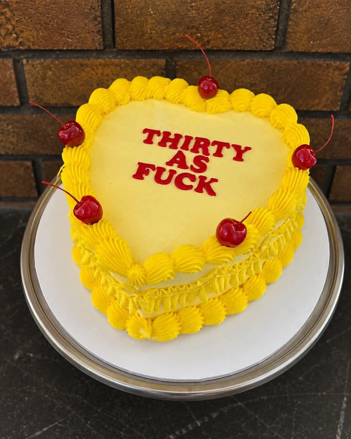 Yellow heart cake with red fondant letters reading Thirty As F*** and red cherries