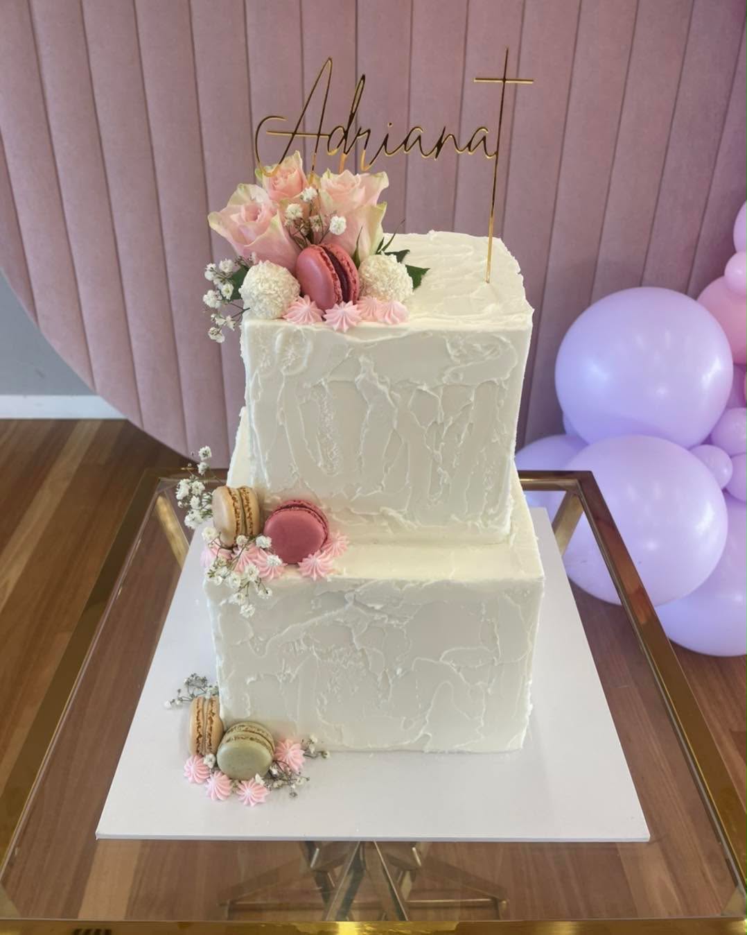 Two-tier square white textured buttercream wedding cake with Adriana gold script topper, pink roses, macarons, white truffles and baby's breath