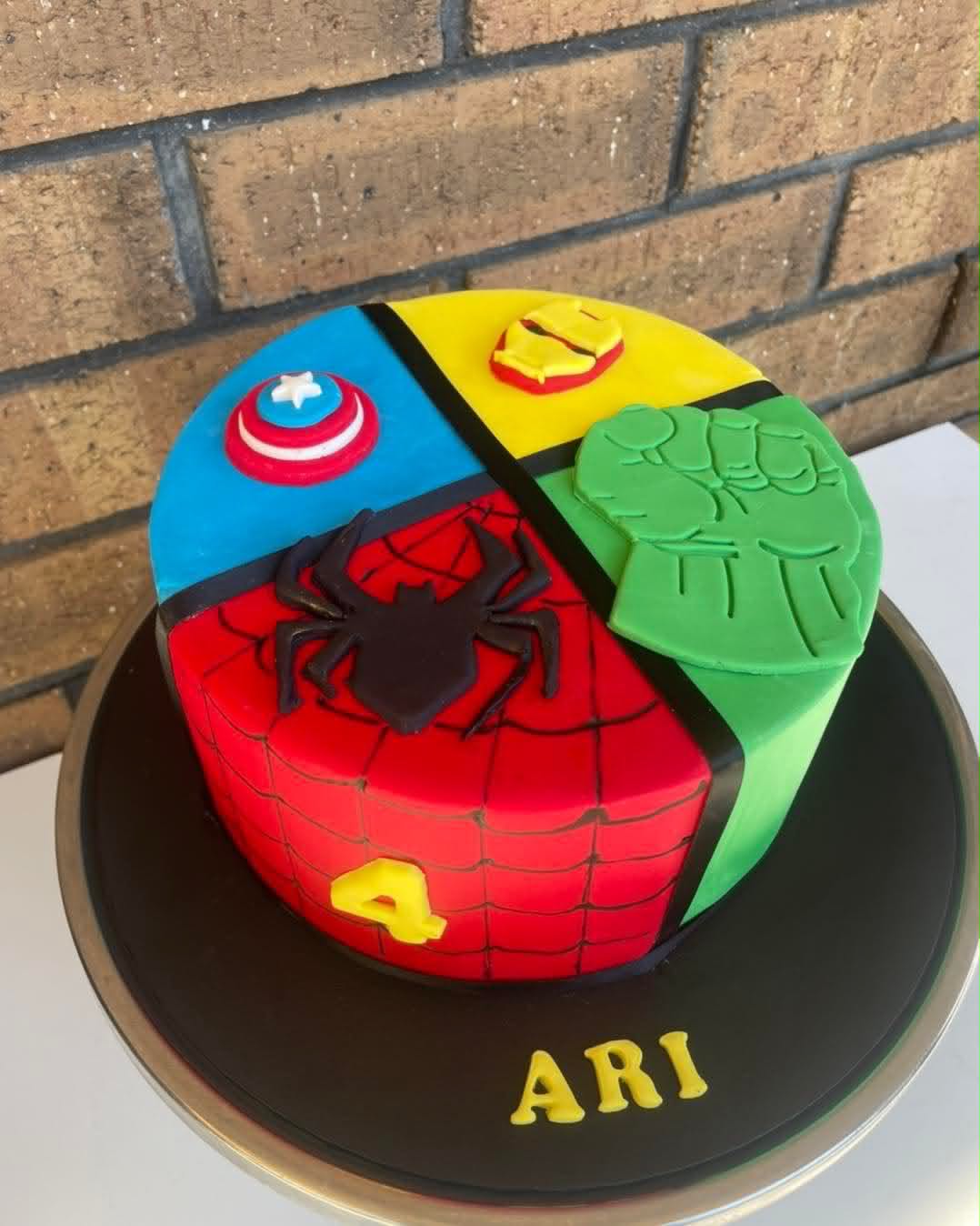 Round Avengers birthday cake for Ari's 4th birthday with four quadrants — blue Captain America shield, yellow Iron Man mask, red Spiderman web with black spider, and green Hulk fist