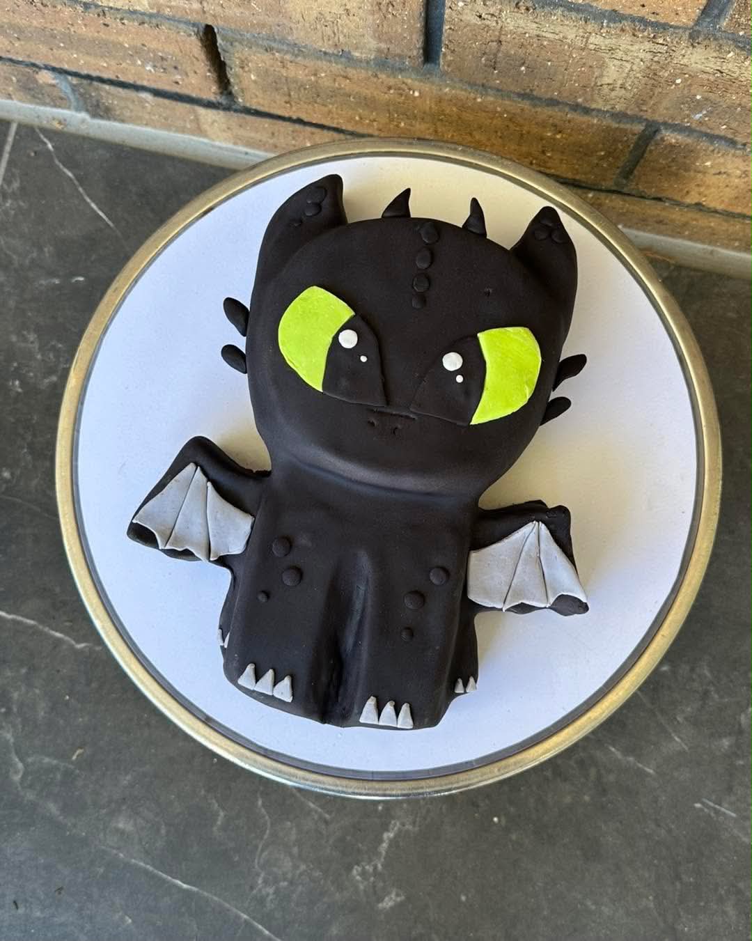 Black How to Train Your Dragon Toothless Night Fury sculpted fondant cake with green eyes, grey wings and white teeth on a silver cake stand