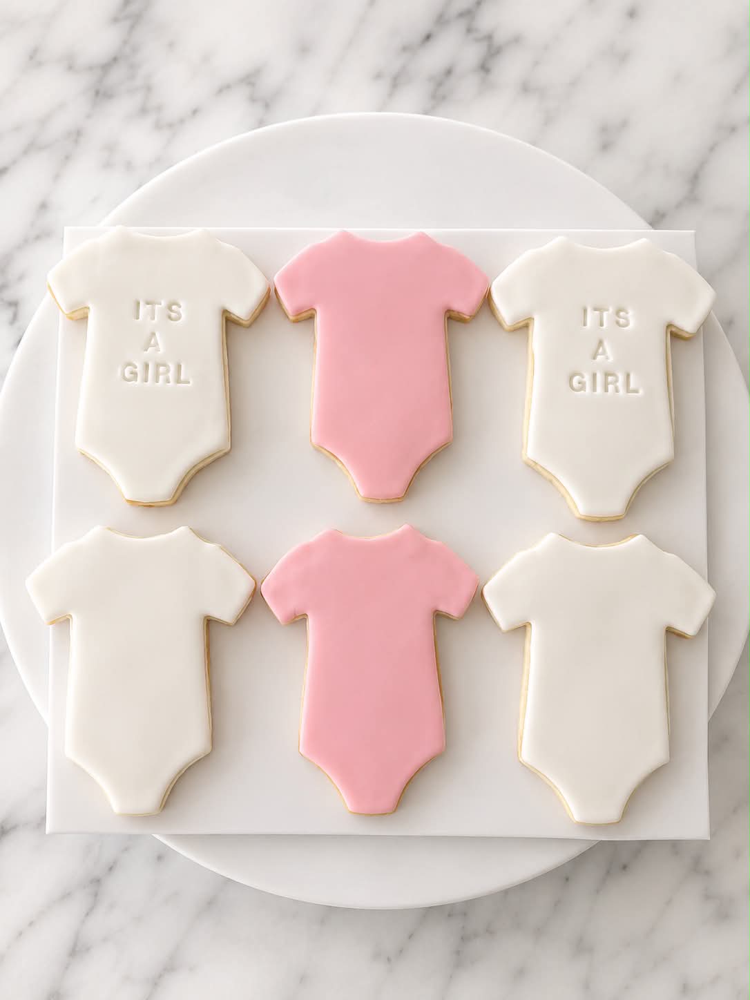 Six white and pink It's A Girl onesie shaped sugar cookies on a white marble board