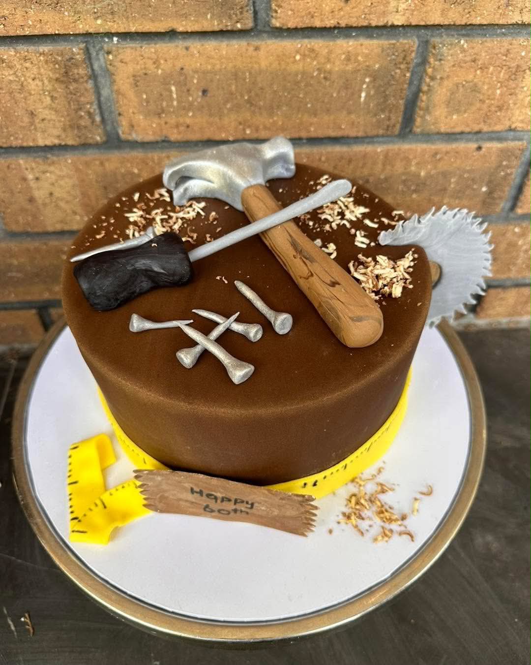 Brown fondant toolbox birthday cake for 60th with fondant hammer, saw blade, nails, chisel and wood shavings on top, yellow tape measure around base and Happy 60th plaque