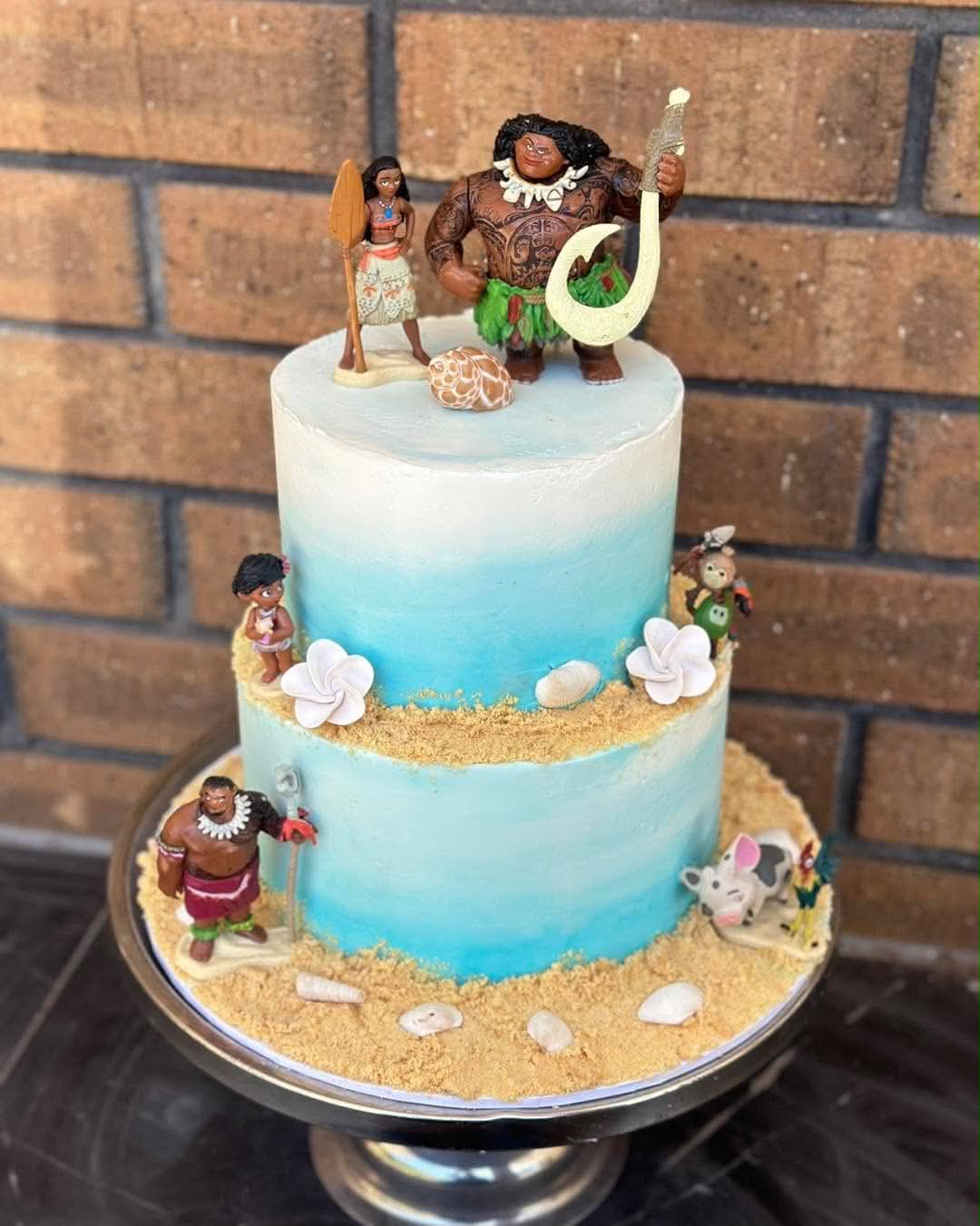 Two-tier blue ombre Moana birthday cake with Moana, Maui, baby Moana, Tamatoa and Hei Hei figurines, sand base, shells and plumeria flowers