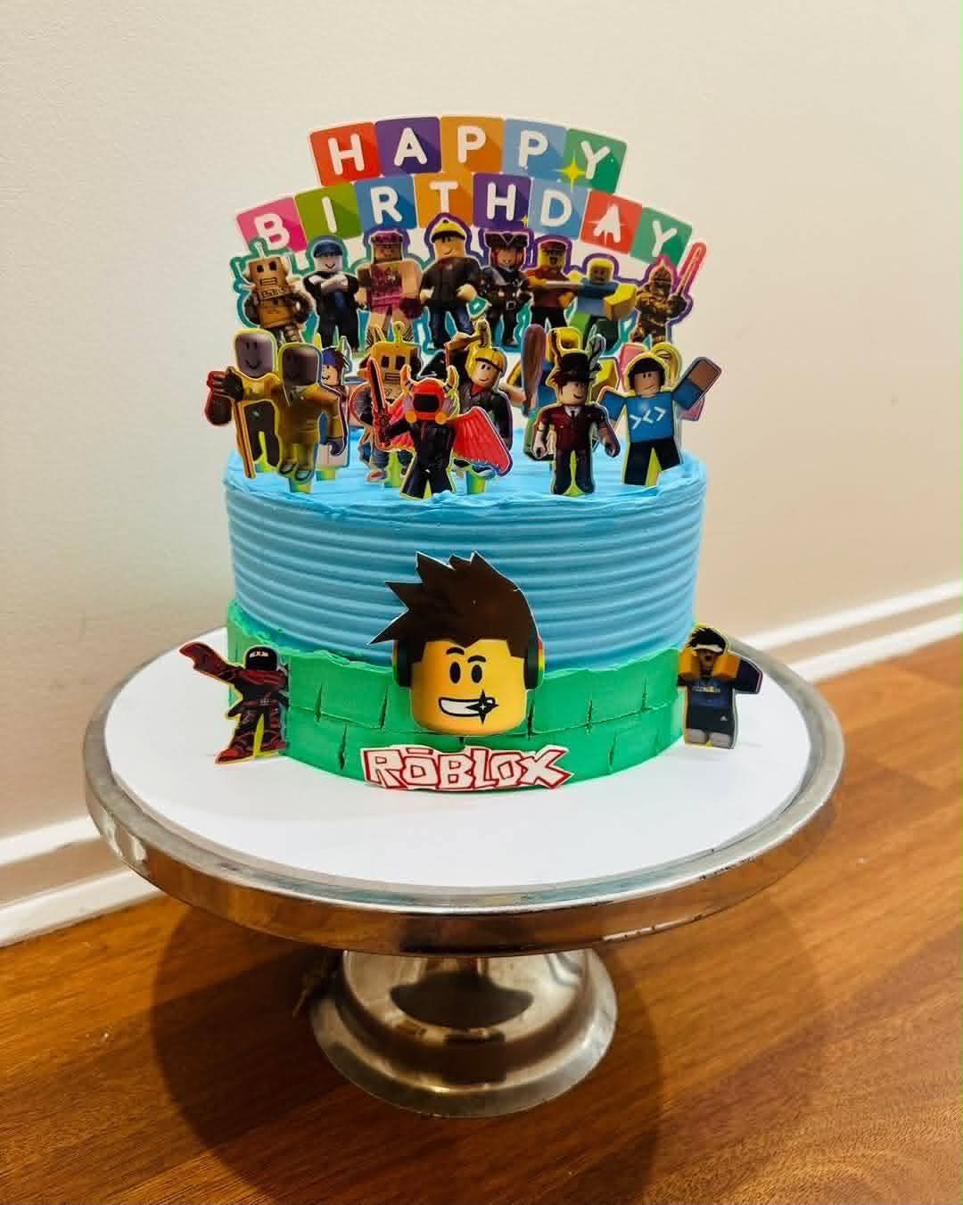 Two-tier blue and green Roblox birthday cake with Roblox character edible prints and Happy Birthday letter block topper