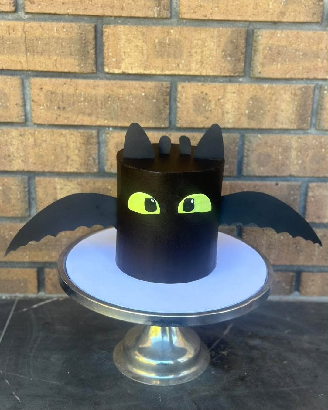 Black Toothless How to Train Your Dragon cake with fondant fin ears, green glowing eyes and black wafer paper wings on silver cake stand