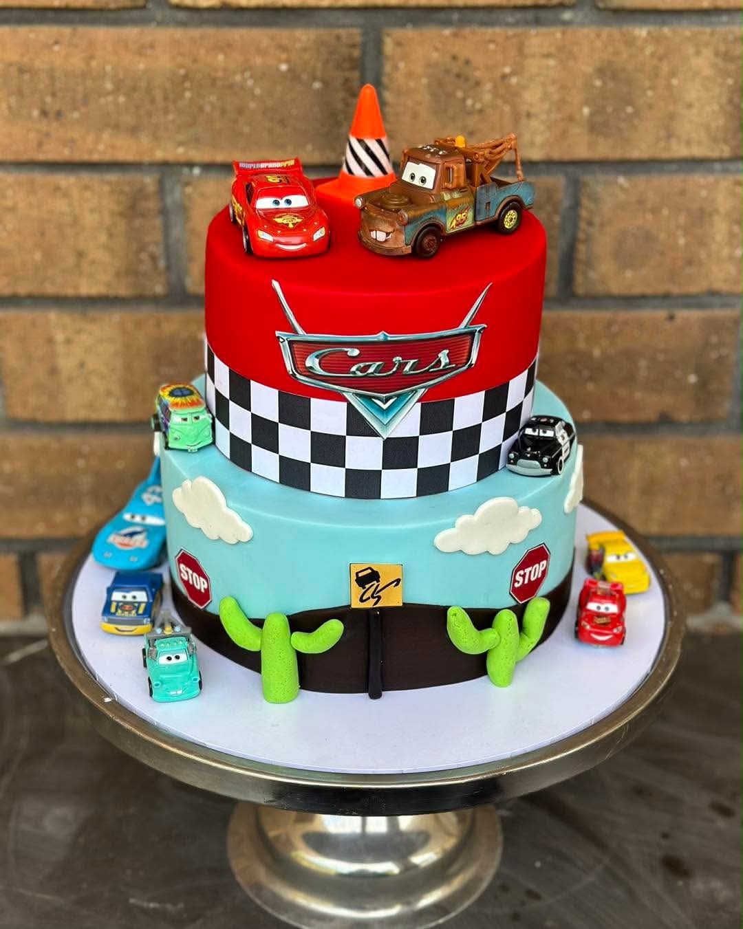 Two-tier Disney Cars birthday cake with red fondant top tier, Cars logo, checkered border, Lightning McQueen and Mater toppers and car figurines