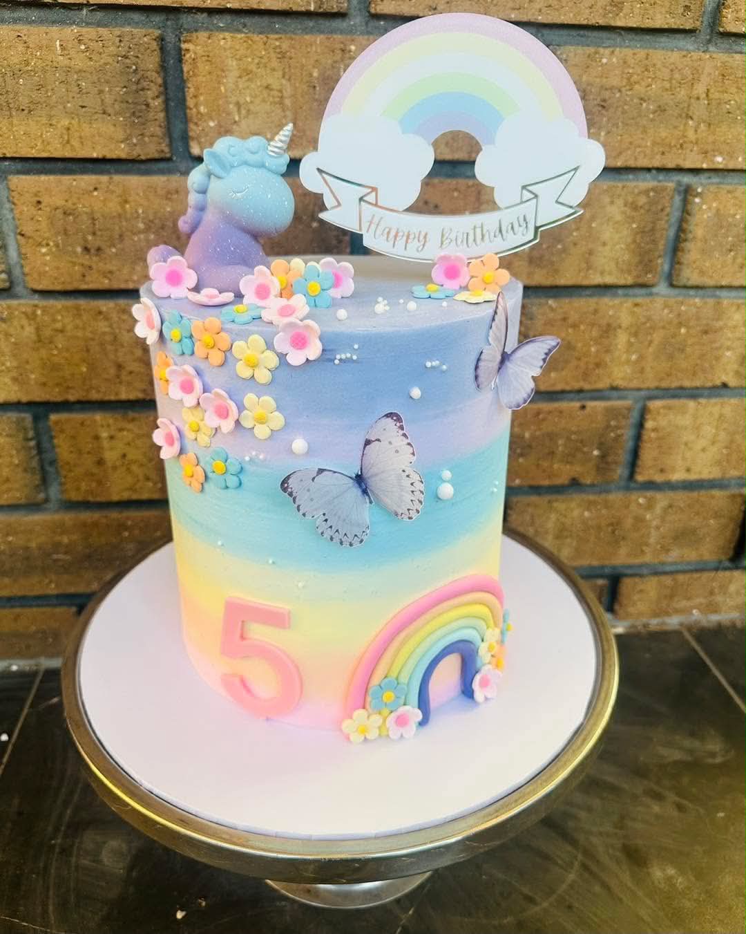 Pastel rainbow unicorn 5th birthday cake with blue unicorn figurine, fondant flowers, butterflies and rainbow arch topper