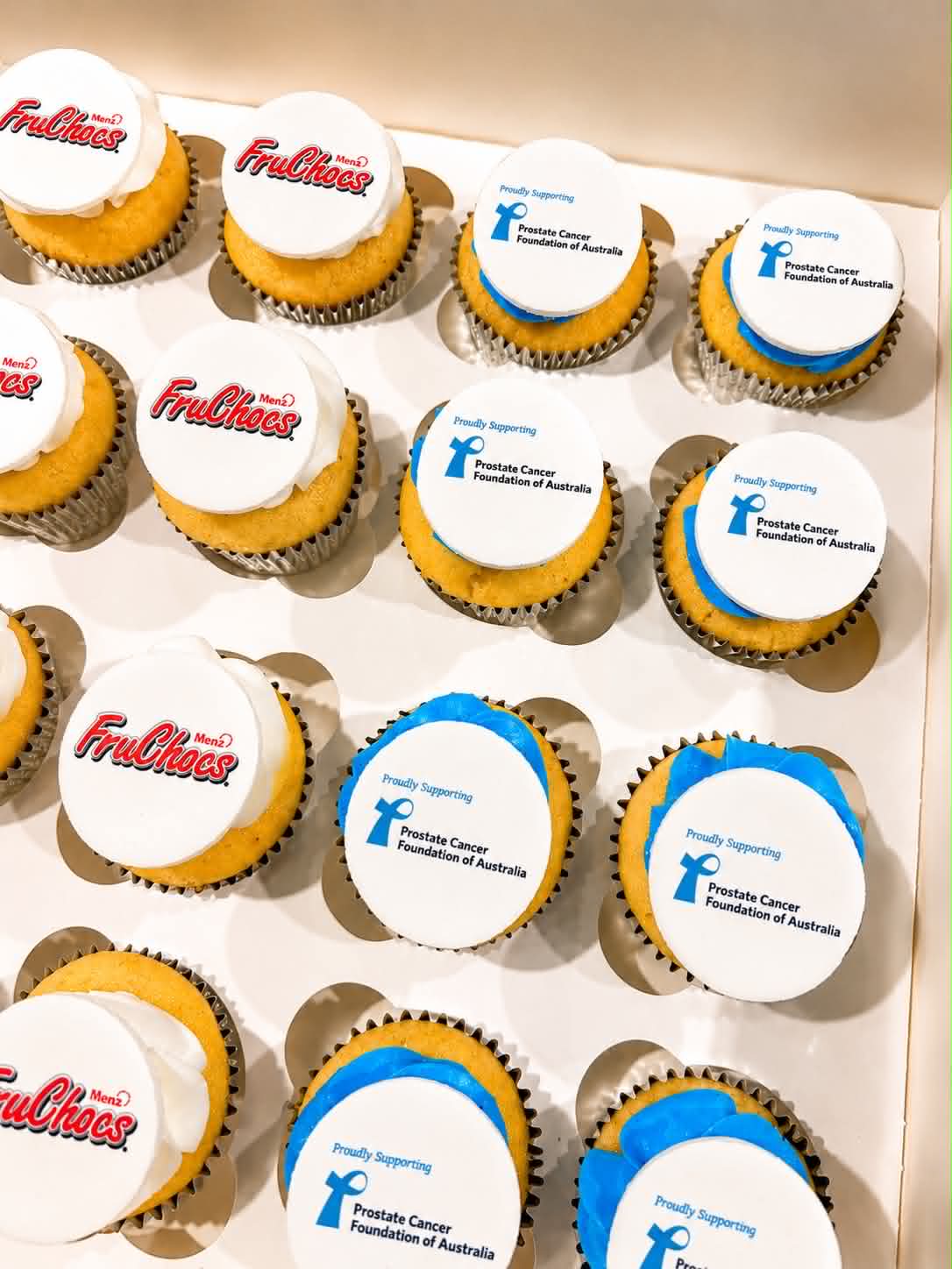 Box of vanilla cupcakes with white fondant toppers — half with FruChocs Menz red logo edible prints, half with blue Prostate Cancer Foundation of Australia Proudly Supporting edible prints