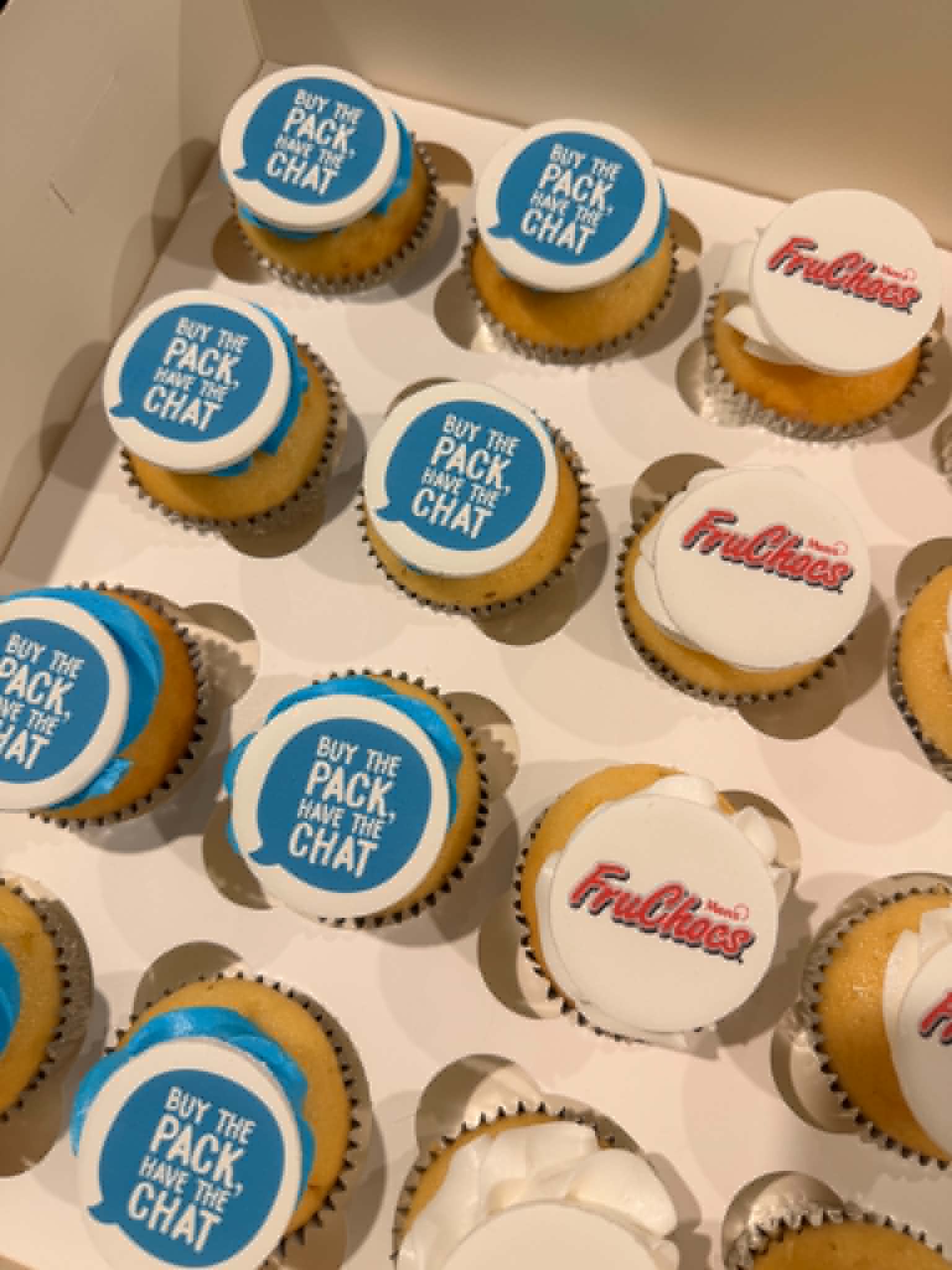 Box of vanilla cupcakes with white fondant toppers — half with blue Buy the Pack Have the Chat speech bubble edible prints, half with FruChocs Menz red logo edible prints