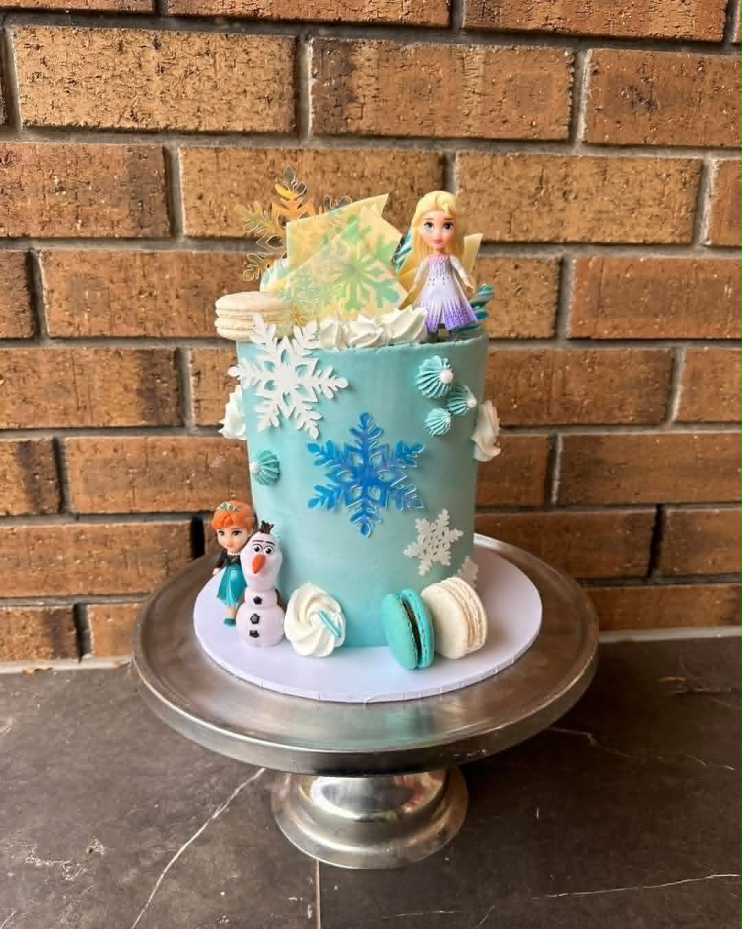 Teal Frozen birthday cake with white and blue snowflake decorations, Elsa figurine on top, Anna and Olaf figurines at the base, macarons and iridescent shards