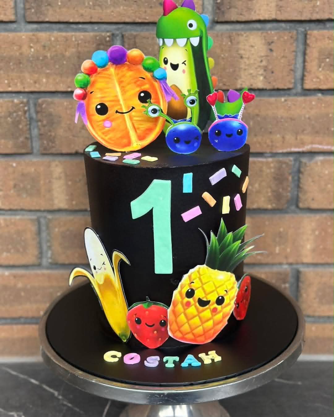 Black two-tier 1st birthday cake for Costah with kawaii fruit and vegetable edible print characters — orange, avocado, blueberries, banana, strawberry and pineapple
