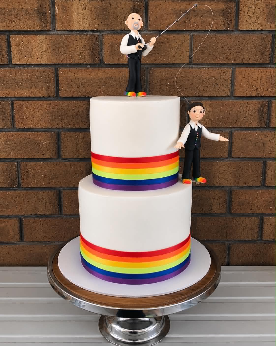 Two tier white fondant wedding cake with rainbow pride ribbon borders on each tier, a fondant groom figurine fishing from the top tier and a second groom figurine on the side