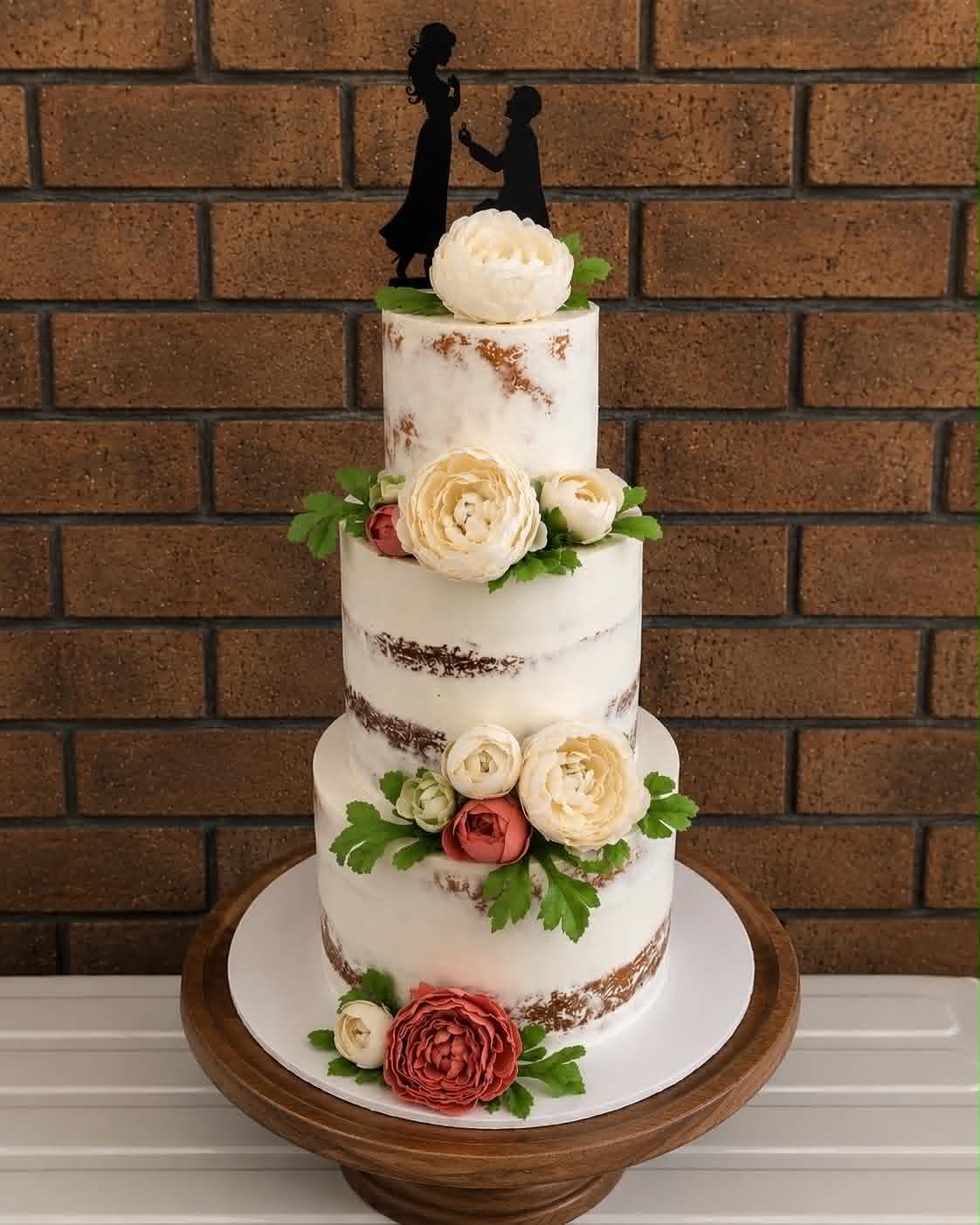 Four tier semi-naked wedding cake with fresh cream and coral peonies, ranunculus and greenery cascading down the tiers and a black proposal silhouette acrylic topper
