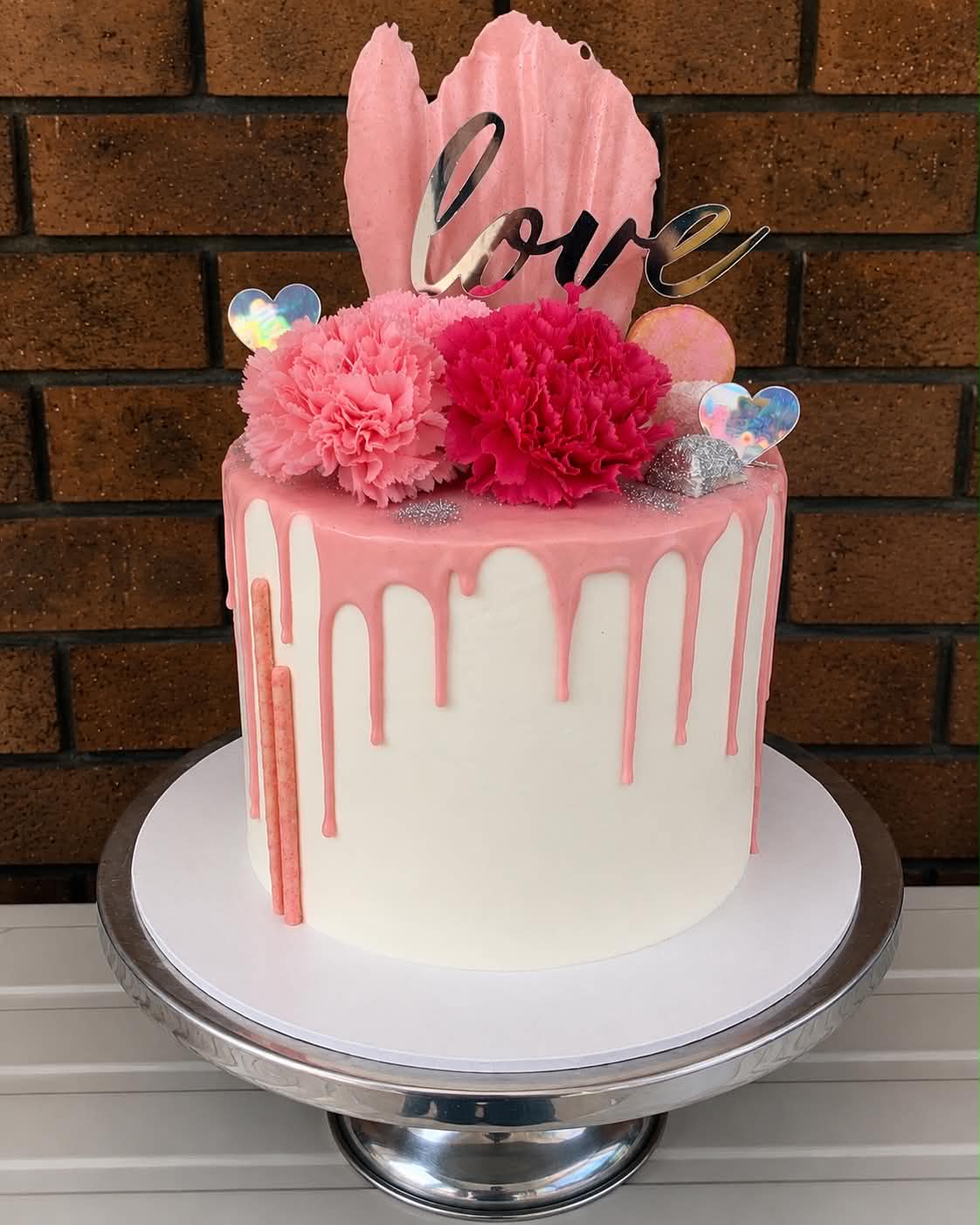 Single tier white buttercream cake with pink chocolate drip, fresh pink and hot pink carnations, holographic heart decorations, pink wafer sail and a silver Love acrylic topper