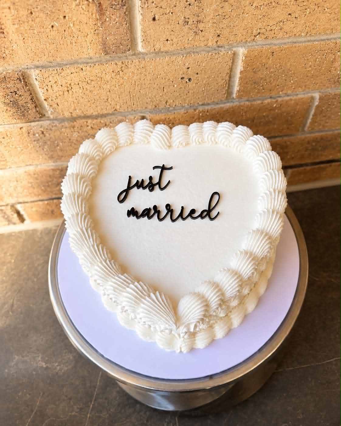 White heart shaped wedding cake with shell and fan buttercream border piping and black acrylic Just Married script topper on a silver cake stand