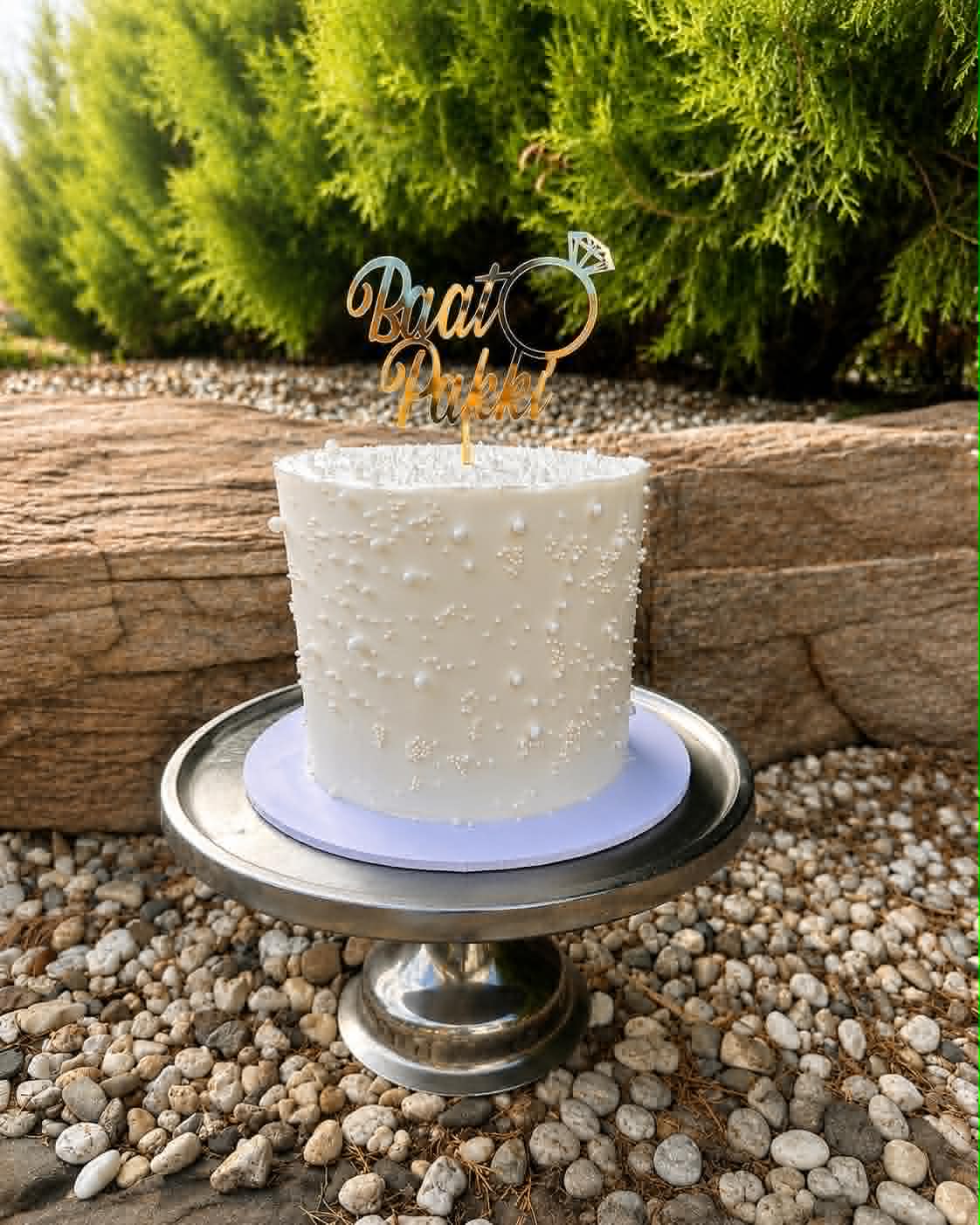 Single tier white buttercream engagement cake covered in white pearl sprinkles with a gold Baat Pakki acrylic ring topper, displayed outdoors on a silver cake stand