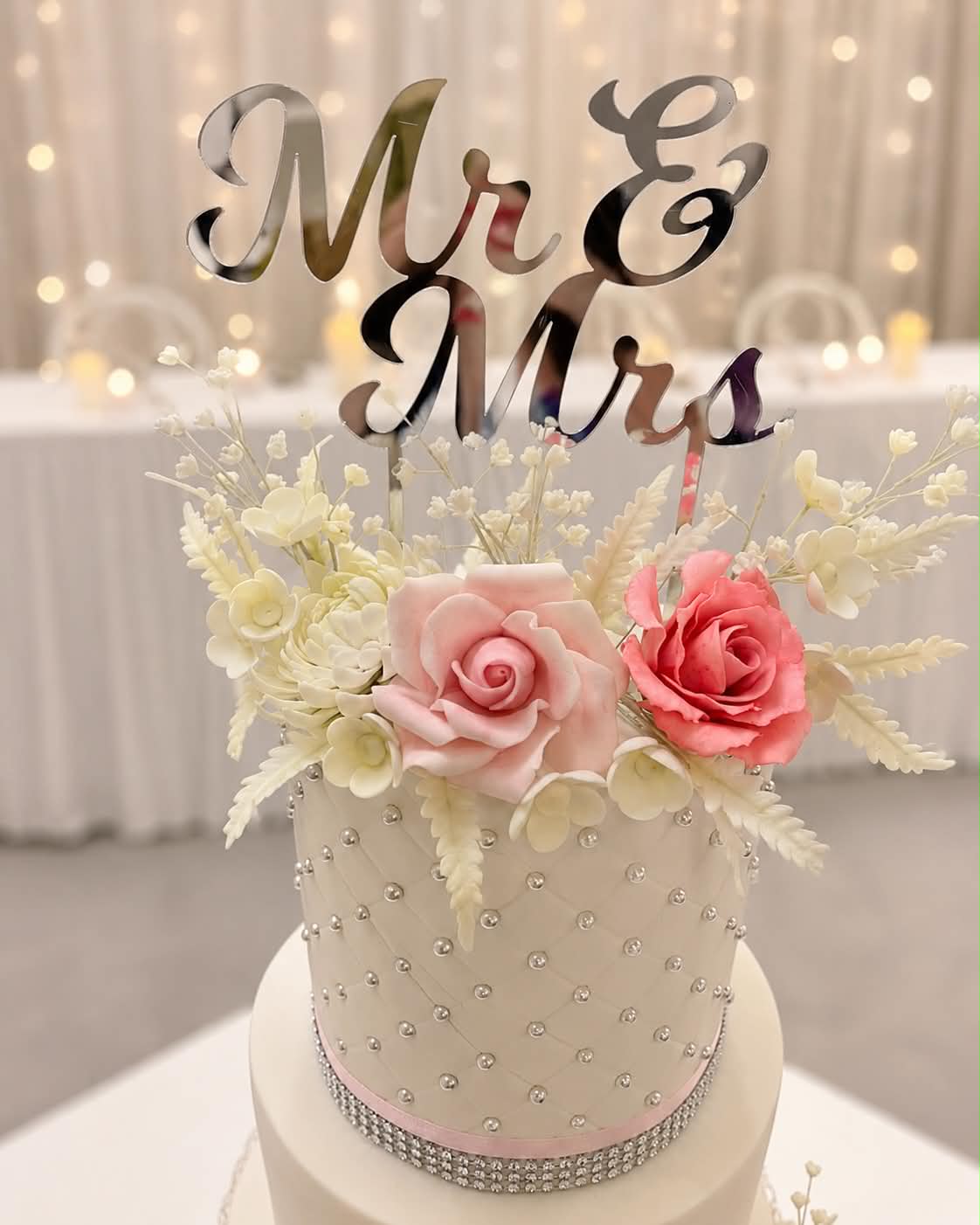 Close up of white quilted fondant wedding cake tier with silver pearl dot grid, pink and coral sugar roses, white gypsophila and fern sprigs and a silver Mr & Mrs acrylic topper