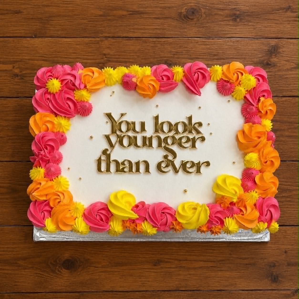 White slab cake with hot pink, orange and yellow buttercream rosette border and You Look Younger Than Ever gold acrylic topper on wooden background