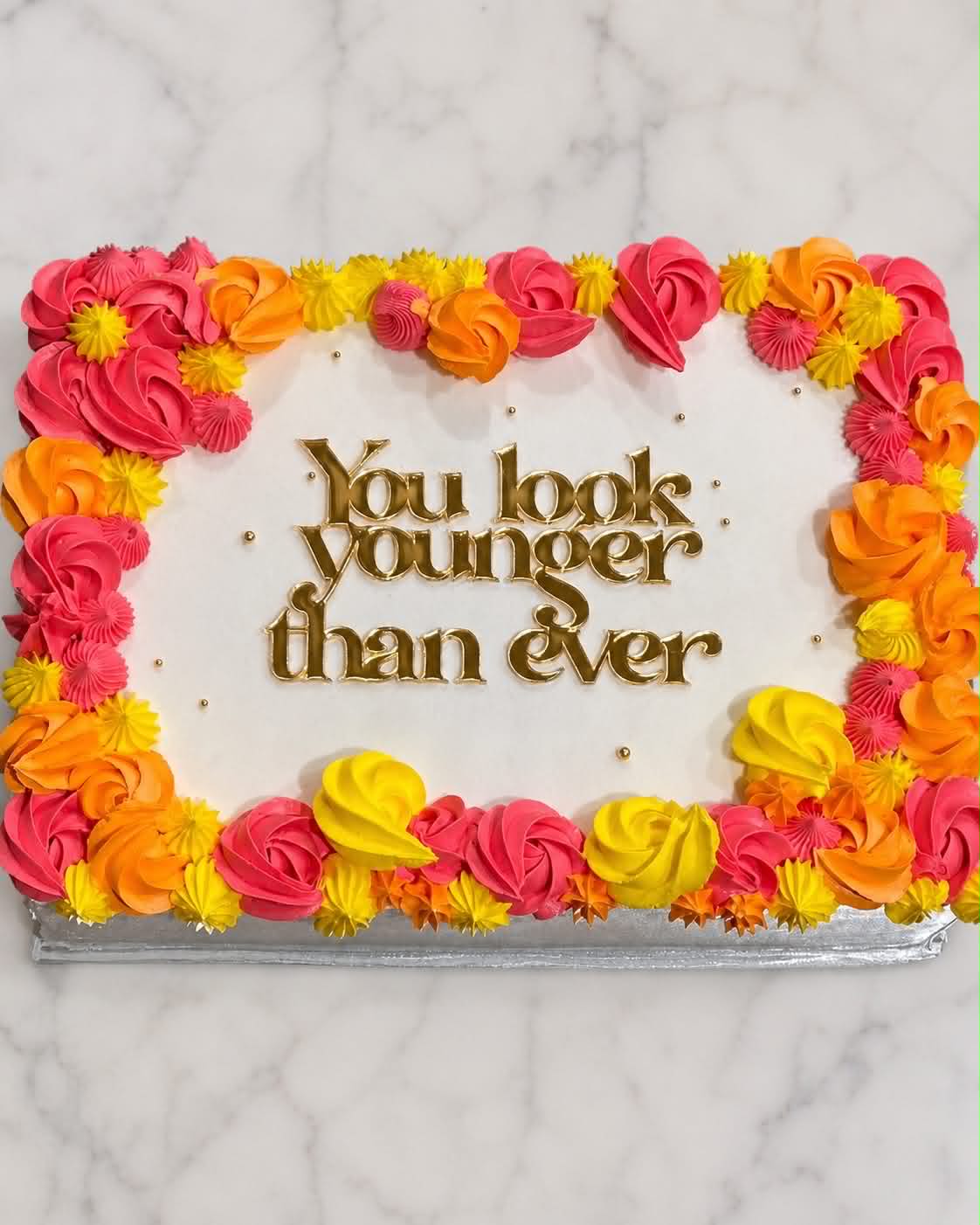 White slab cake with hot pink, orange and yellow buttercream rosette border and You Look Younger Than Ever gold acrylic topper on marble background