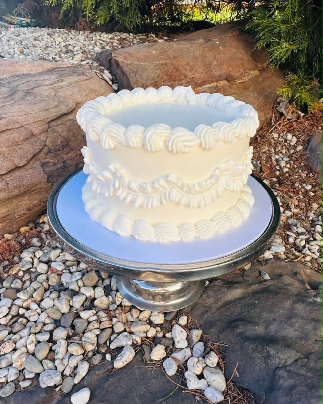 Ivory buttercream vintage cake with shell and scroll piping border on top and middle, displayed on a silver cake stand outdoors on pebbles