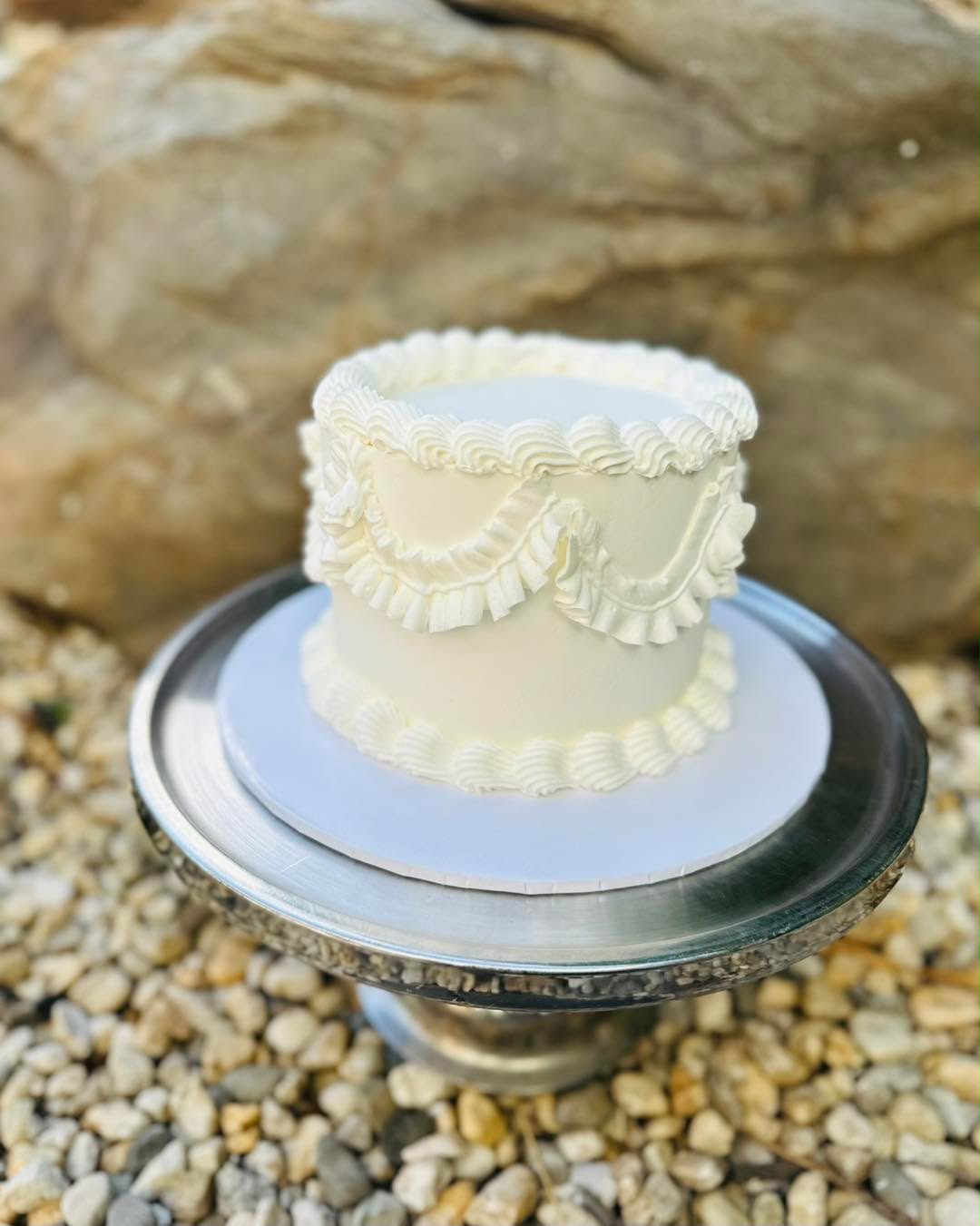 Ivory buttercream vintage cake with ruffle and frill piping and swag detail on a silver cake stand outdoors on pebbles
