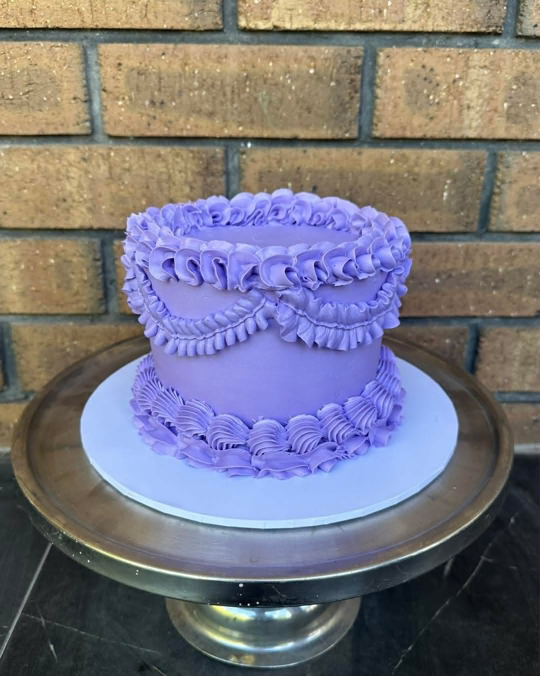 Lilac purple buttercream vintage cake with ruffle and frill piping and swag detail on a silver cake stand against a brick wall