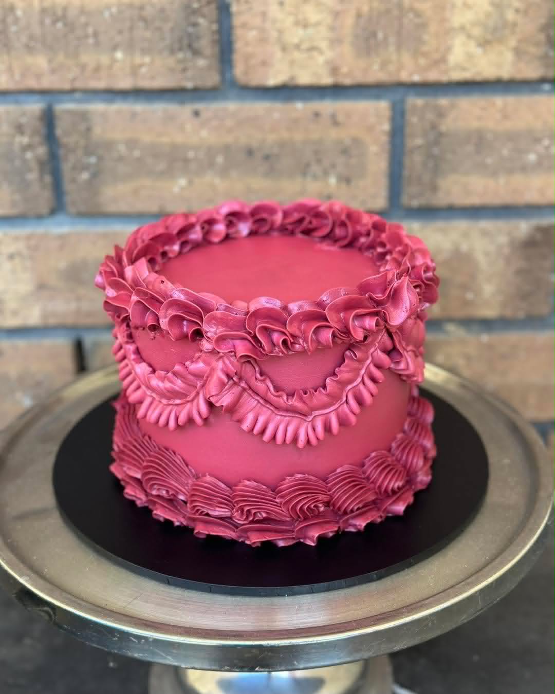 Deep crimson red buttercream vintage cake with ruffle and frill piping and swag detail on a silver cake stand against a brick wall