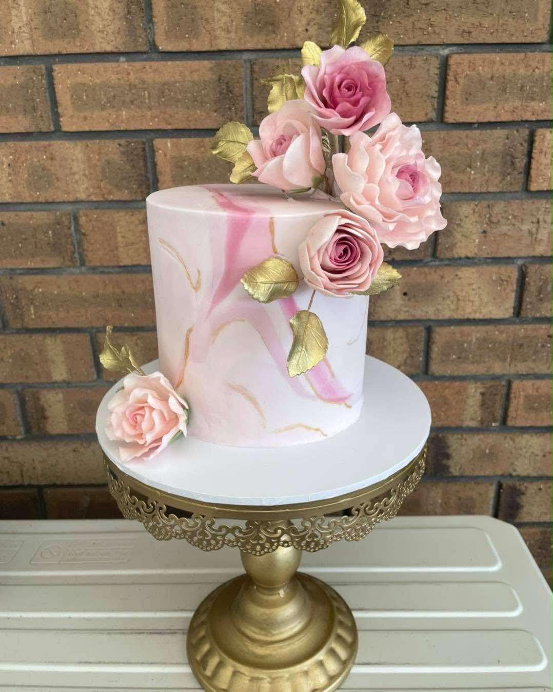 Single tier white fondant wedding cake with pink and gold marble effect and large pink sugar roses and gold leaves on a gold filigree cake stand