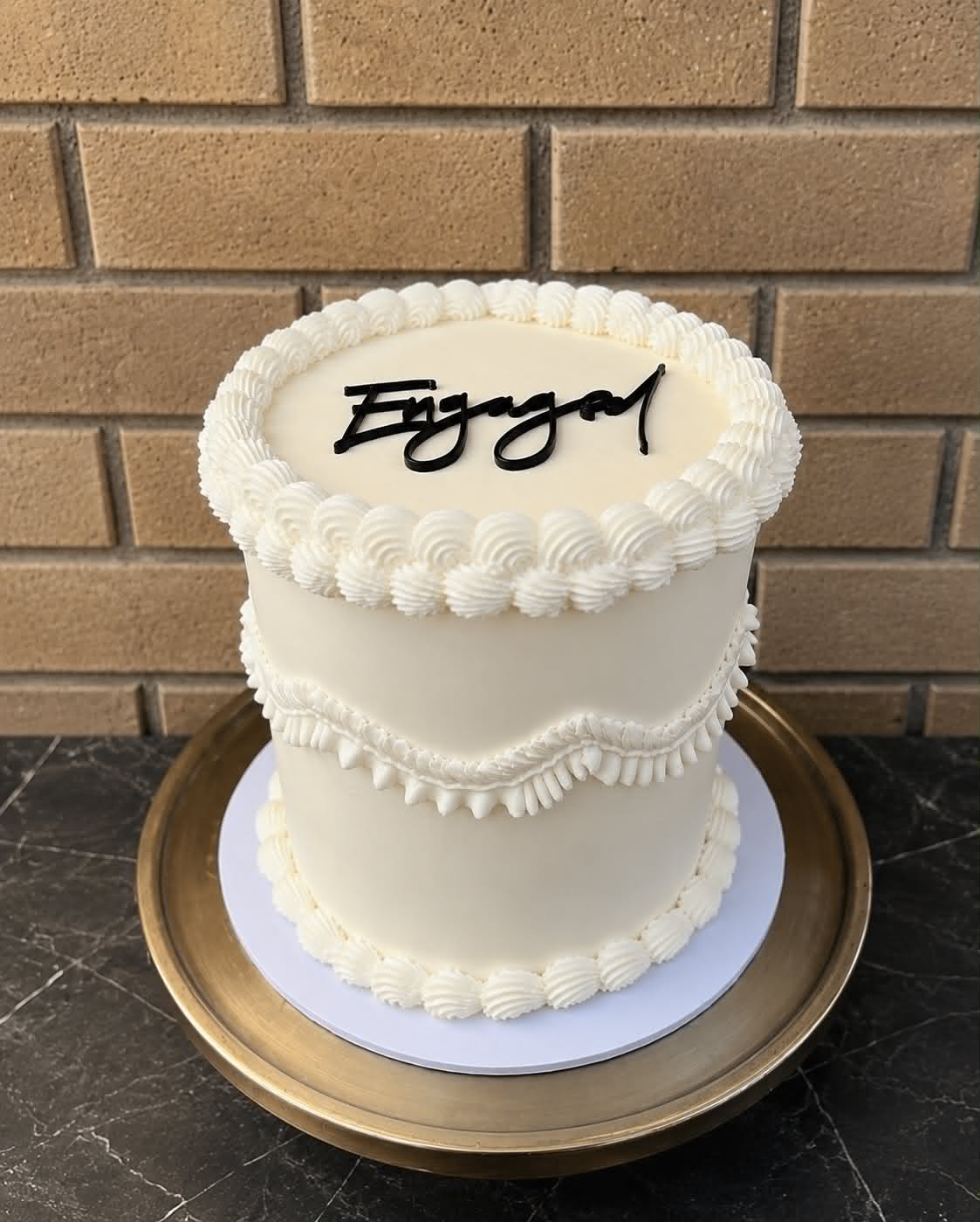 Ivory buttercream engagement cake with rosette and shell piping border and black acrylic Engaged script topper on a gold cake board